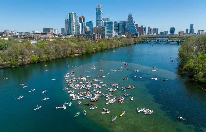 Colorful kayaks and paddleboards on blue-green water with a view of the Austin skyline in the background.