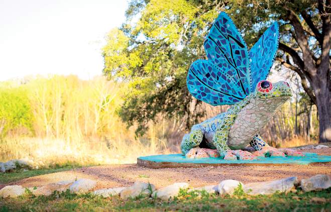 Colorful mosaic sculpture of a frog with a large blue frill, set against a blurred natural background with trees and grass.