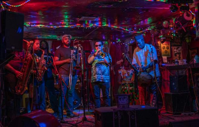 Photo of 5 men in a band on stage at the Sahara Lounge. The room is dark and rainbow rope lights surround the ceiling. From left to right person playing baritone sax, man playing alto sax, man holding trombone, man playing trumpet, man playing kora