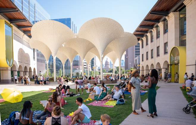 Groups of people standing around and sitting on picnic blankets across the Blanton Museum's lawn, in between museum buildings.