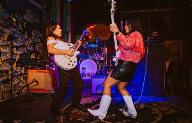 Two Latina women playing guitars on a stage with colorful lights.
