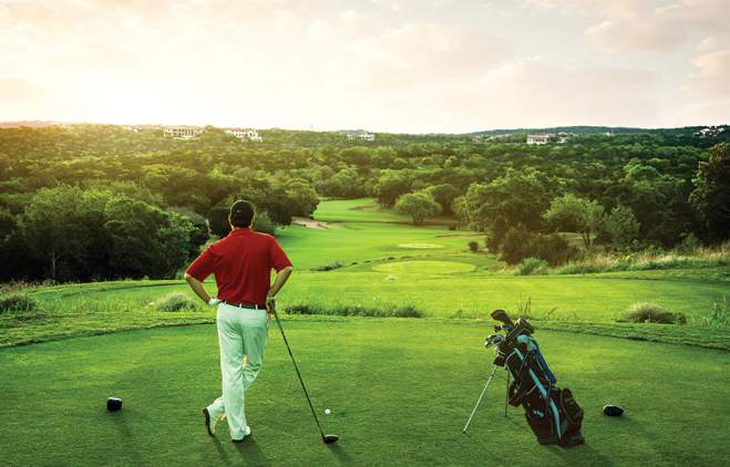 Male golfer stands on a teebox with driver and bag looking out towards a sunset