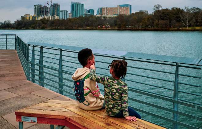 Two young children sitting on a bench on the Hike & Bike Trail Boardwalk. Lady Bird Lake and the Austin skyline are behind them