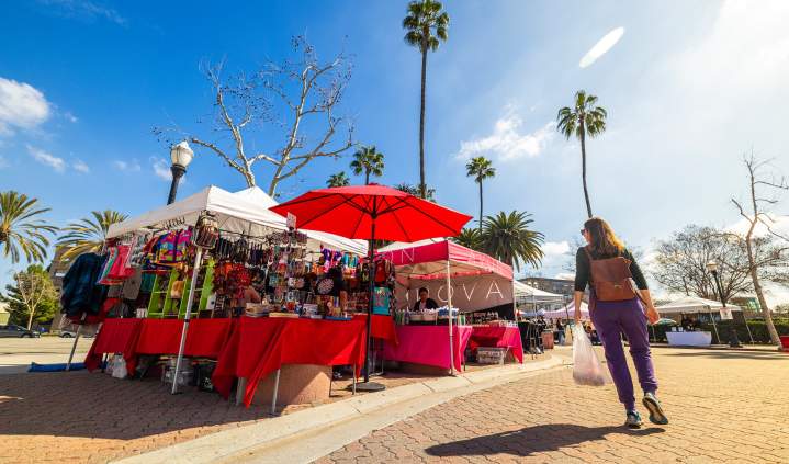 Anaheim Farmers Market in Orange County, California, featuring colorful vendor booths with red umbrellas, shoppers browsing local goods, and palm trees surrounding an outdoor plaza on a sunny day.