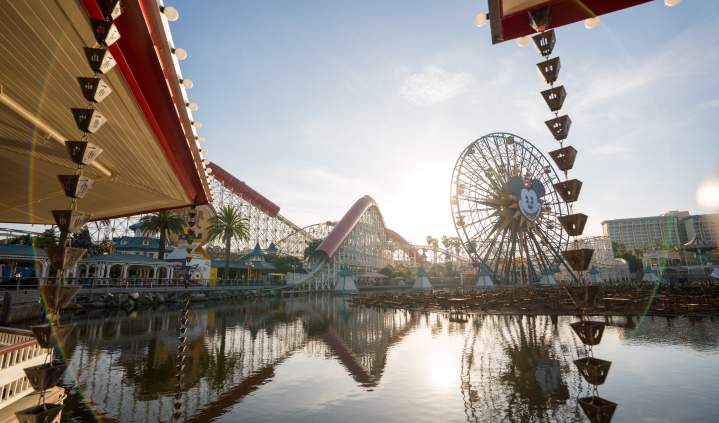 Sunrise at Disney California Adventure Park with a large Ferris wheel and roller coaster, reflected in a serene lake surrounded by palm trees.