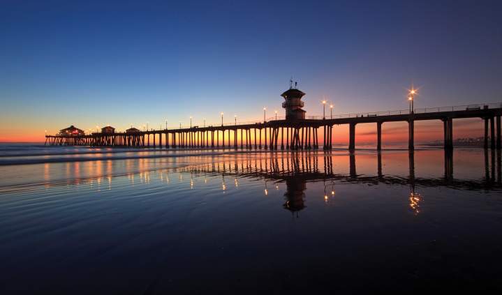 Huntington Beach pier at sunset with lights reflecting on wet sand, extending into calm ocean waters under a gradient sky of deep blue and warm orange hues.