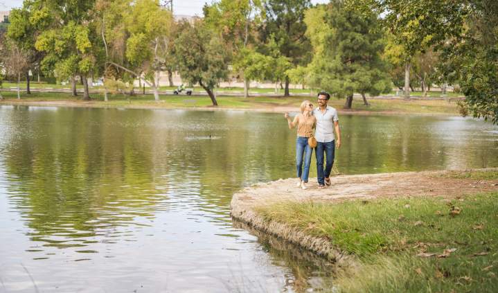 Yorba Regional Park in Anaheim featuring two people walking along a grassy shoreline beside a calm lake, with trees, open green space, and gentle ripples on the water under a bright sky.
