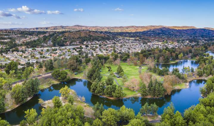 Yorba Regional Park in Anaheim featuring a wide aerial view of scenic lakes, tree-lined green spaces, walking paths, and nearby neighborhoods with rolling hills in the distance under a clear blue sky.