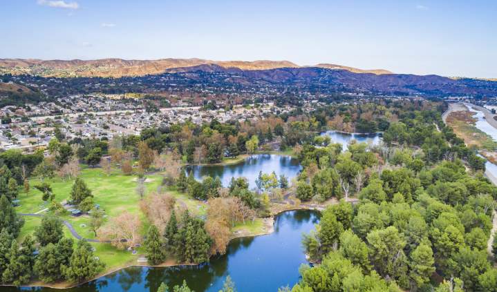 Yorba Regional Park in Anaheim featuring an aerial view of tree-lined lakes, winding walking paths, open green spaces, and surrounding residential neighborhoods under a clear blue sky.