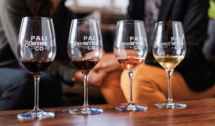 Pali Wine Co. wine tasting flight featuring four glasses of red, rosé, and white wine arranged on a wooden table, with guests seated in the background inside a relaxed tasting room.