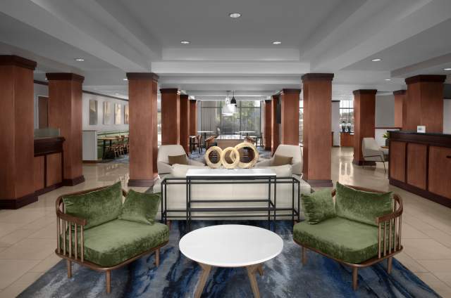 Lobby with cherry wood pillars throughout, a wooden lobby check-in desk to the right and seating in the middle with sandy colored tile floors. The middle of lobby has two velvet green chairs with a small circular side table in the middle, a white entry table with metal framing, and a white couch with two white chairs behind the entry table. A blue modern streaked carpet runs down the middle. In the background a variety of bar stool and cafe-style seating can be seen.