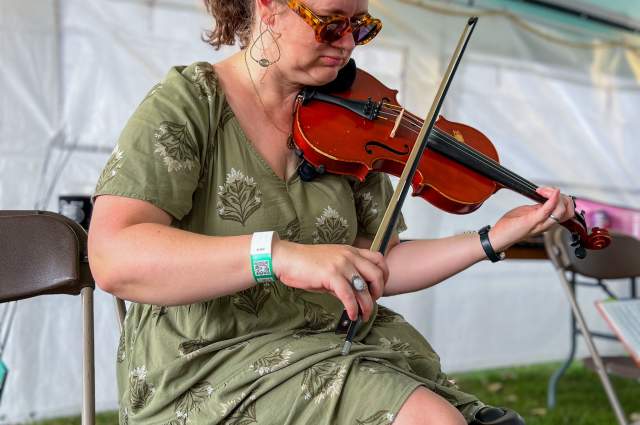 A musician seated on a chair under a tent, playing a violin. They are wearing a green dress and sunglasses, and have a festival wristband on.
