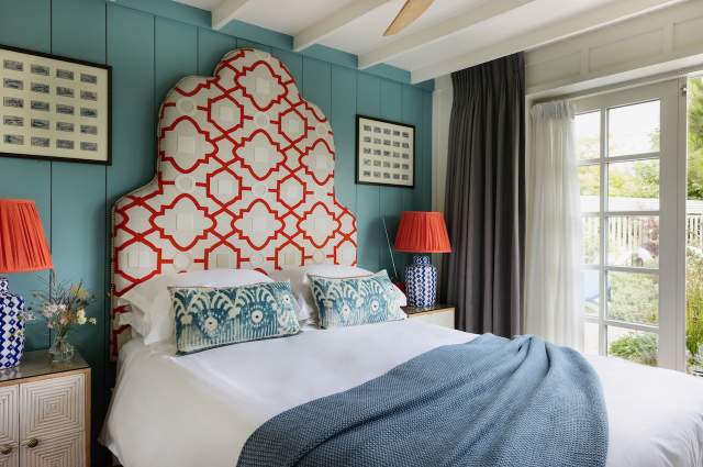 Interior shot of Bedroom at The Gallivant coastal hotel, a bed with white sheets and coloured pillows and a dark blue throw. The headboard is tall with red detail and there are red bedside lamps. Light streams through a patio door from the garden.