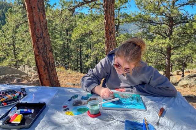 A young woman paints on a small canvas at a table set up in a Ponderosa pine forest
