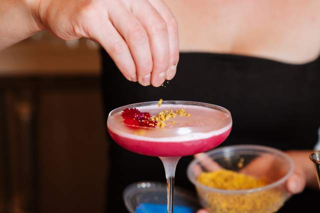 A mixologist tops a bright pink cocktail off with a pinch of yellow bee pollen