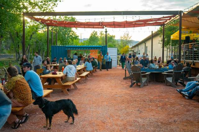Outdoor patio at Pattern Break Brewing in Boulder with picnic tables, string lights, guests enjoying drinks, and a dog at sunset