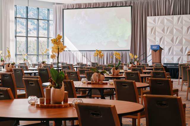 Limelight Boulder Flatirons Ballroom set in rounds for a conference, featuring floor-to-ceiling windows with mountain views, a presentation stage and screen, and elegant orchid centerpieces