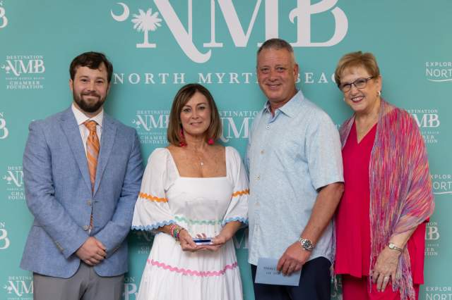 Four individuals pose together in front of a teal "Destination North Myrtle Beach" backdrop. From left to right: a man in a light blue blazer and orange-striped tie; a woman in a white dress with colorful trim, holding a clear award; a man in a light blue short-sleeve shirt holding an envelope; and a woman in a vibrant pink dress with a multicolor shawl. All are smiling and appear to be at a formal recognition or award event.