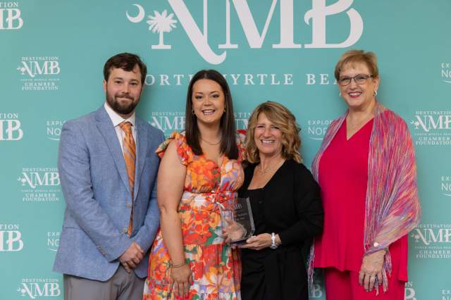 Four individuals pose in front of a teal "Destination North Myrtle Beach" backdrop. From left to right: a man in a light blue blazer and orange-striped tie; a woman in a colorful orange and pink floral dress with her arm around the woman next to her; a woman in black holding a clear award with text visible on it; and a woman in a bright pink outfit with a multicolor shawl. All are smiling and appear to be part of an award or recognition ceremony.