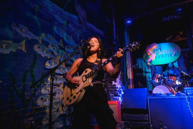 Woman standing on a stage painted with an irredescent fish backdrop, playing the electric guitar and singing into a microphone.