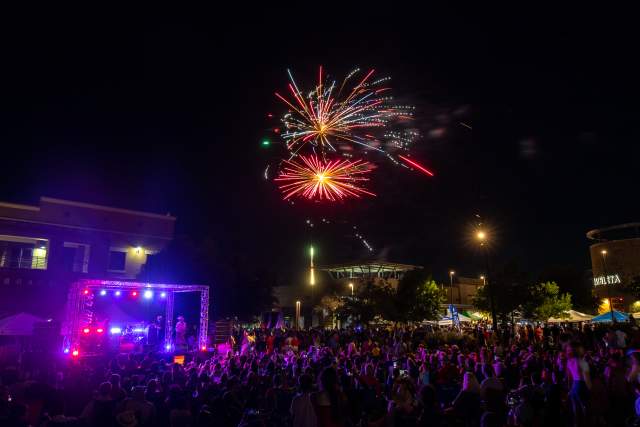 Fireworks exploding in the dark sky above a crowd of people and a band onstage at the Hill Country Galleria Independence Day Festival.