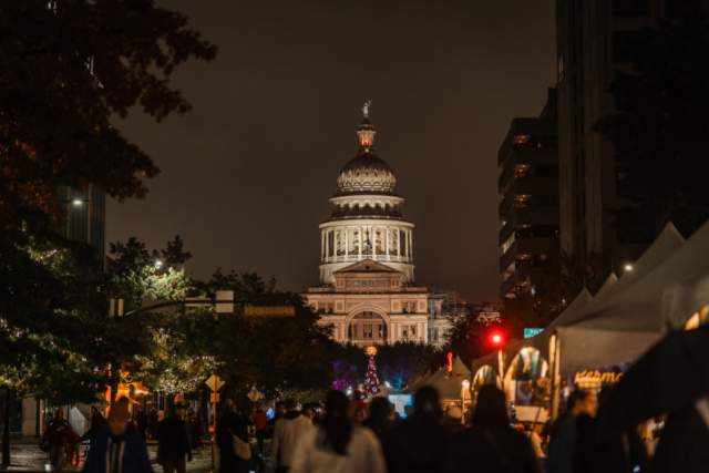 The Texas State Capitol Building illuminated at dusk with a group of people standing in the streets in front of the building.