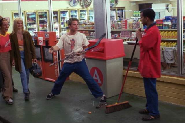 Suburbia screengrab, showing a man in a red shirt sweeping the ground while another man stands in front of him with a hockey stick in his hand. A group of people are walking past them and laughing