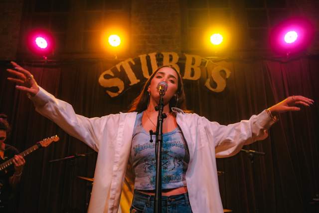 Woman standing in front of a microphone, singing with arms out, a large sign saying "STUBB'S" with pink and yellow neon lights is above her head.