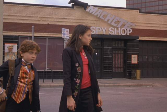 Spy Kids screengrab showing Carmen and Juni walking together in front of a building with sign reading Machete Spy Shop