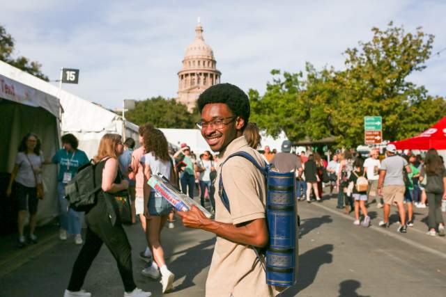 Image of a man holding a book and walking down Congress Avenue while looking over his shoulder to smile at the camera with the Texas State Capitol in the background.