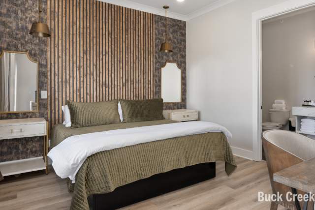 A modern hotel room with a wooden slate wall and green bedding located at Buck Creek BnB. The room has a desk and a bathroom.
