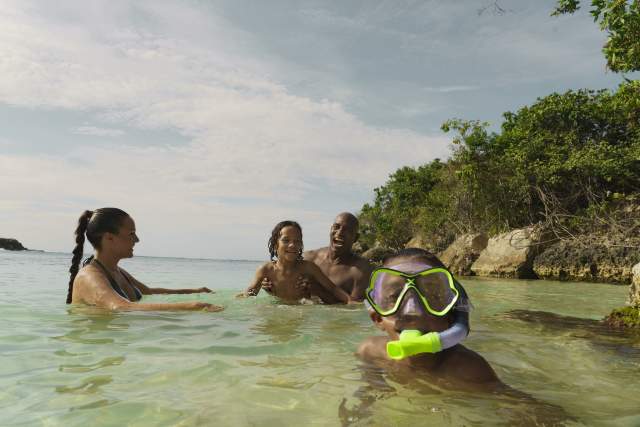 A family plays together in the water at a beach in Jamaica. In the foreground, a little boy with goggles and a snorkle looks at the camera.