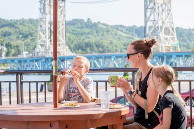 Two children and adult eat at outside picnic table with Portage Lake Lift Bridge in background.