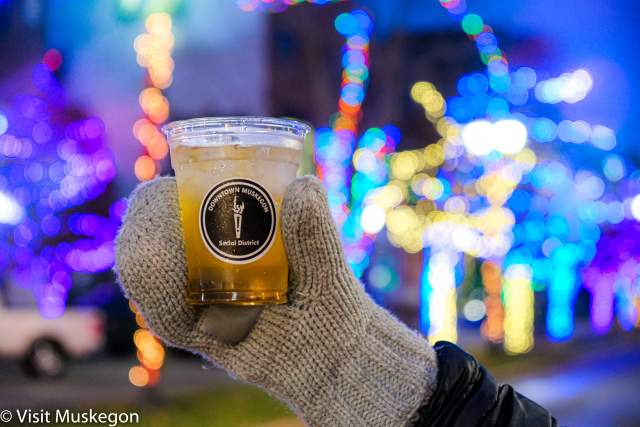 mittened hand holds clear plastic cup filled with drink. front of cup shows downtown muskegon social district label. background is filled with glowing strings of holiday lights