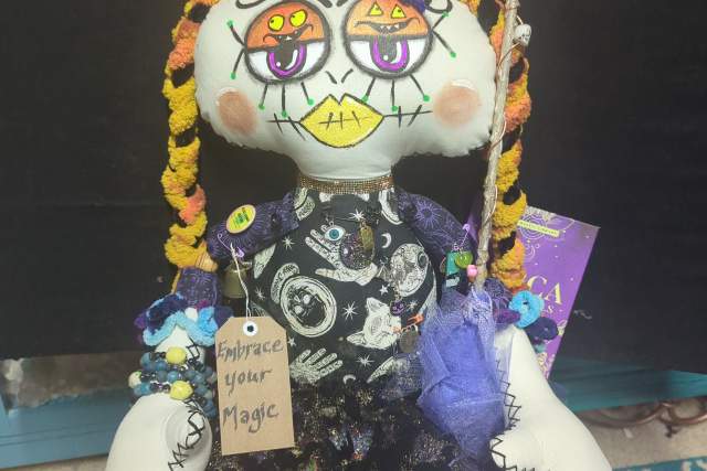 a white, black and purple doll, accented by unique designs sits on a shelf. A tag reads "embrace your magic"