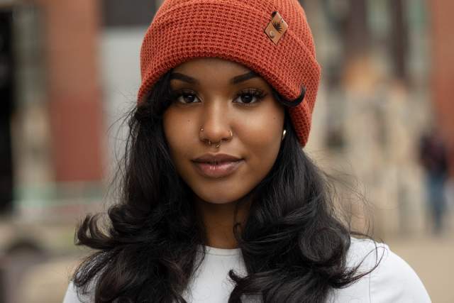 Portrait of a smiling individual wearing a burnt orange beanie, with a city street in the background.