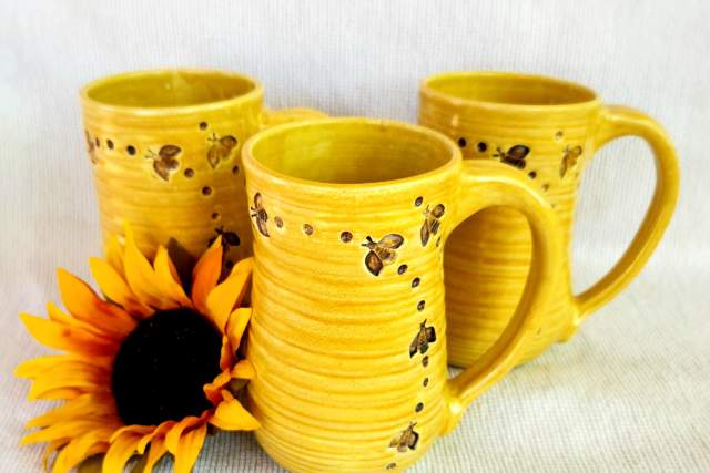 three yellow clay mugs with bees flying around the outside sit on a white backdrop with sunflower beside them