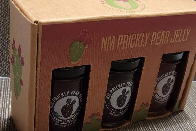 Three jars of jelly are housed in a cardboard container that displays the labels. The box reads "NM Prickly Pear Jelly" and the label reads the same.