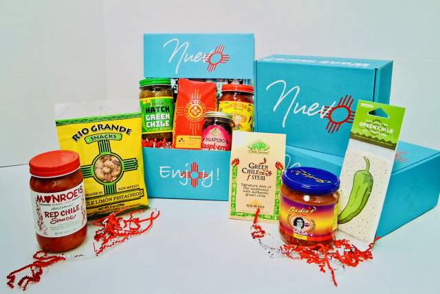 Assorted New Mexico food products displayed, including Monroe's Red Chile Sauce, Hatch Green Chile Salsa, and Green Chile Stew. Items are arranged in front of and around blue Nuevo boxes with a white backdrop.