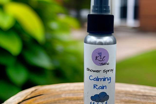 a single silver spray bottle with a black cap and white label sits on a wooden table with an out of focus bush and building behind it. Label reads "KC Naturals, Shower Spray, Calming Rain"