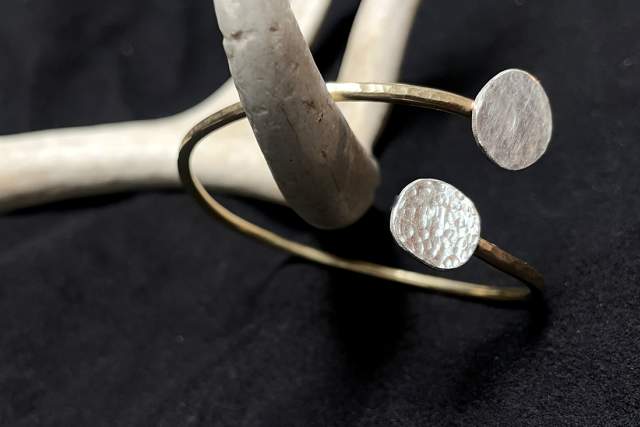 Brass and silver bracelet featuring a plain band with two contrasting textures on circular discs, hanging from a white antler displayed on a textured black surface.
