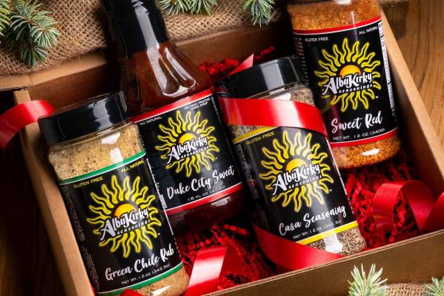A gift box containing four AlbuKirky Seasonings products: Duke City Sweet barbecue sauce, Sweet Red barbecue sauce, Green Chile Rub, and Casa Seasonings, surrounded by festive pine branches.