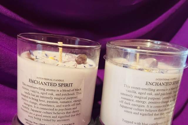 Two "Enchanted Spirit" scented candles on a purple fabric background, each with a detailed label describing the fragrance notes and topped with small stones.
