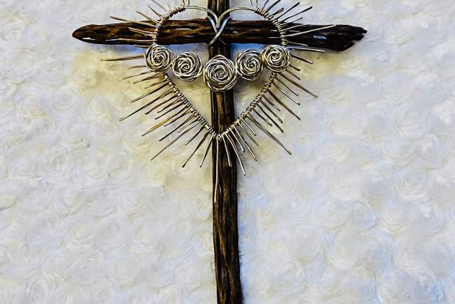 A wooden cross adorned with a heart made of twisted wire and spiral designs is placed on a textured, white surface. The heart is surrounded by a halo of wire spikes.