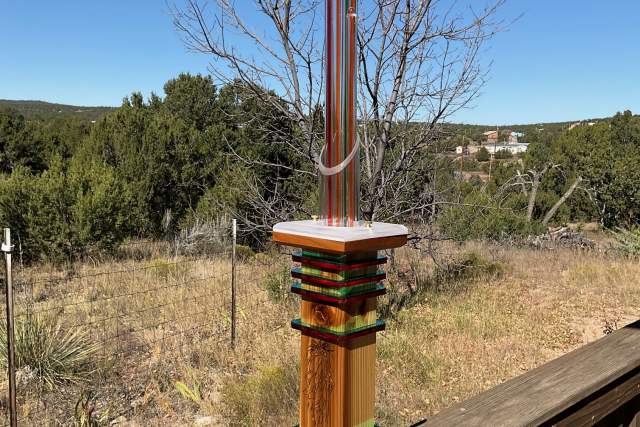 A colorful wooden sculpture with different length lightbulbs protruding from the top, displayed on a wooden banister in front of a field and trees.
