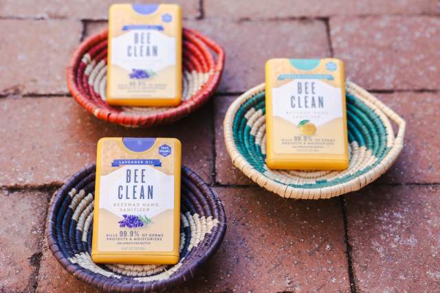 Three "Bee Clean" beeswax hand sanitizers displayed on colorful woven baskets on a brick surface.