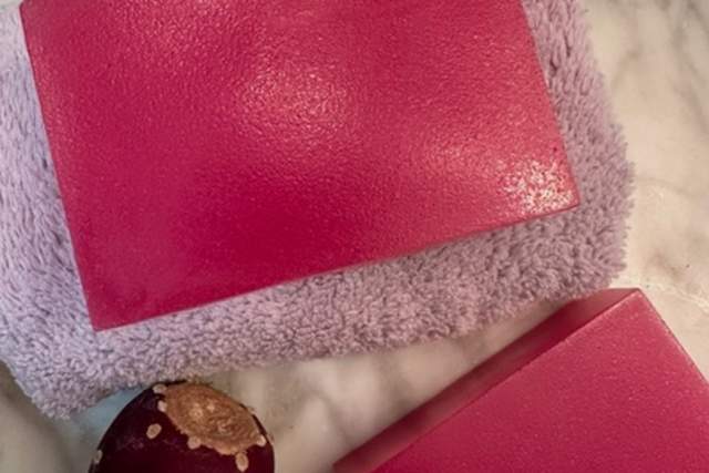 Three pink rectangular soap bars are placed on a soft lavender towel. Nearby, there's a single prickly pear fruit. The background is a white marble surface.