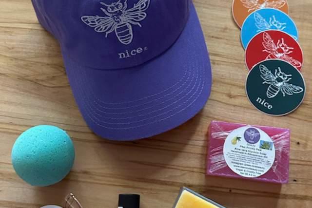 Various personal care products and a purple baseball cap with a bee logo and the word "nice" on a wooden surface. Products include bath bombs, soap bars, and essential oils.