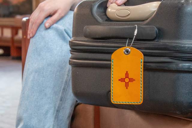 A person sits on a chair with a black suitcase. The suitcase has an orange tag displaying the New Mexico flag symbol.