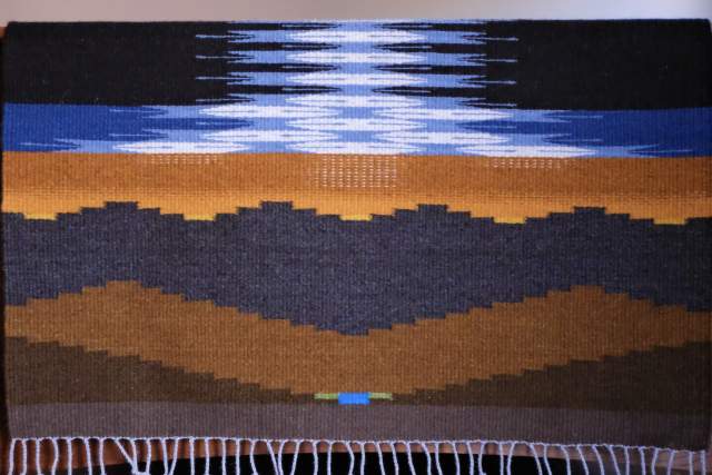 Woven tapestry featuring a landscape with layered colors representing earth and sky, topped by a rippling water effect.